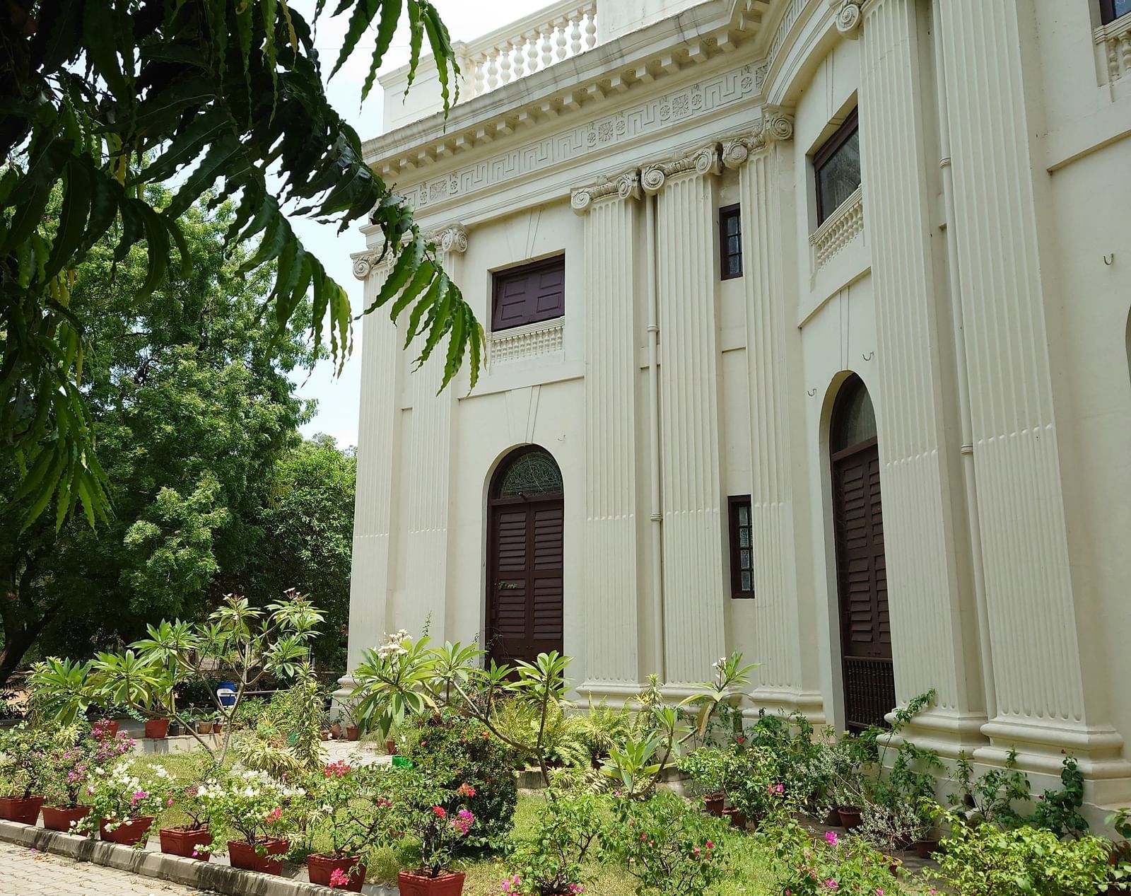 A white heritage-style building with columns and arched windows, surrounded by plants and trees.