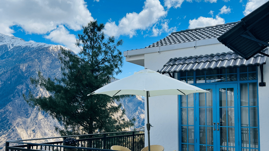 A white cottage featuring a black railing, a white sun umbrella, and a small seating area overlooking a mountain landscape under a bright blue sky at Faraway Cottages, the best hotel in Auli.