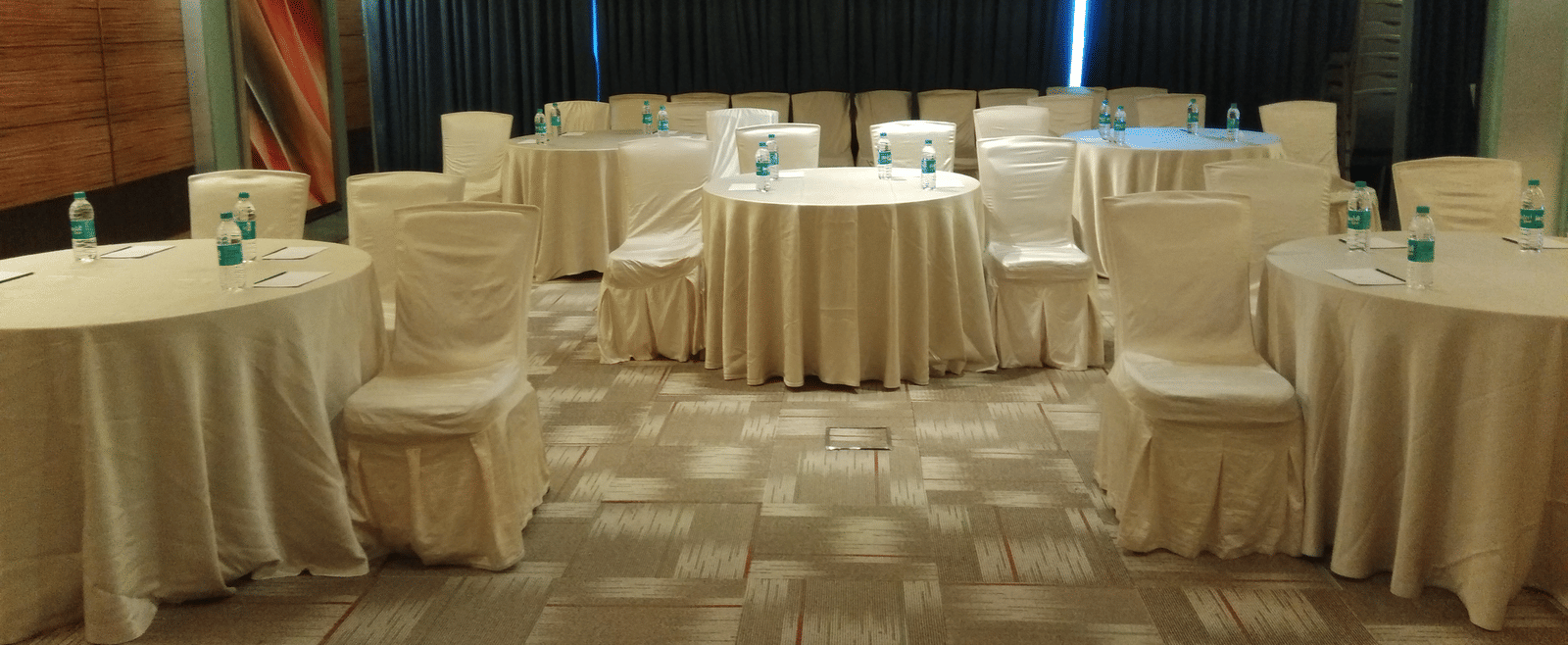 Banquet hall at Mirage Hotel, Mumbai featuring ample seating area, round tables covered in white silk covers and carpet flooring.