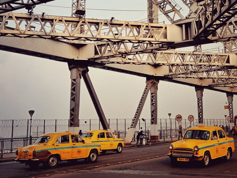 Howrah Bridge structure with steel beams overhead and yellow taxis passing along a busy Kolkata roadway