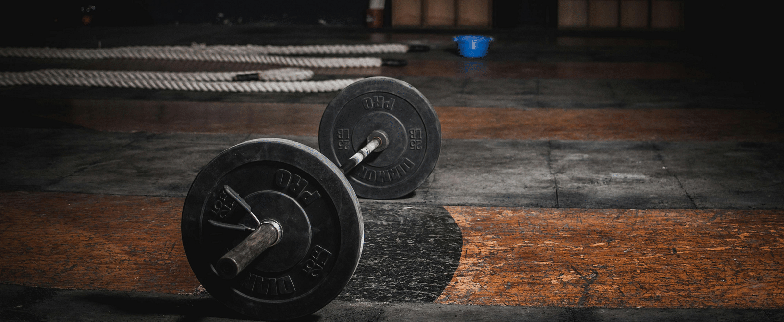 A barbell with weights lies on a worn gym floor, with long metal bars in the background.