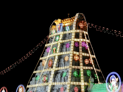 A tall, brightly lit Hindu temple tower (gopuram) stands out against the dark night sky.