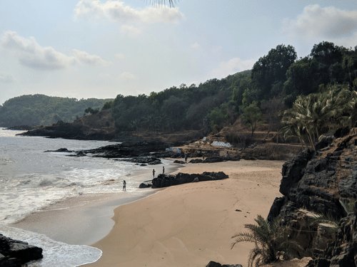 A quiet sandy beach curves beside rugged rocks and swaying palms, framed by gentle hills and the tranquil shoreline beyond.
