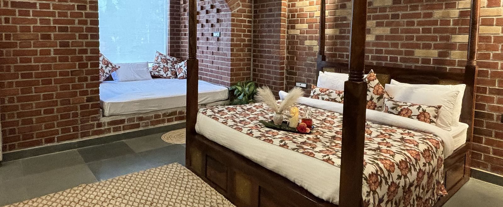 Double bed with brick wall backdrop and arched window in a cosy room at Tisya Farms, Gurgaon.