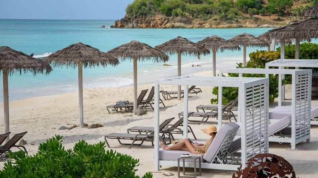 A woman reads on a lounge chair on a white sand beach with turquoise water and thatched umbrellas.