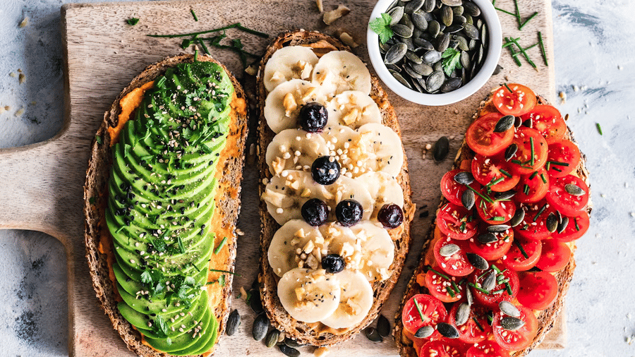 3 open toasts on a wooden board topped with sliced avocado, banana with blueberries, and cherry tomatoes, beside a bowl of pumpkin seeds.