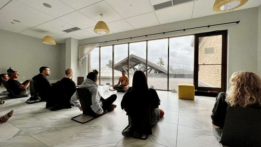 A group of people sits on the floor in a bright, modern room with large windows overlooking an outdoor area and facing the instructor in front at YO1 Longevity & Health Resorts