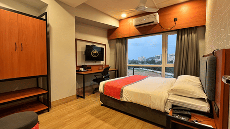 A stylish guest room at Citi Residenci Hotel, Palashdiha, featuring a comfortable double bed, an air conditioning unit above a large window, a sleek wooden wardrobe, and a dedicated desk setup under a wall-mounted TV.