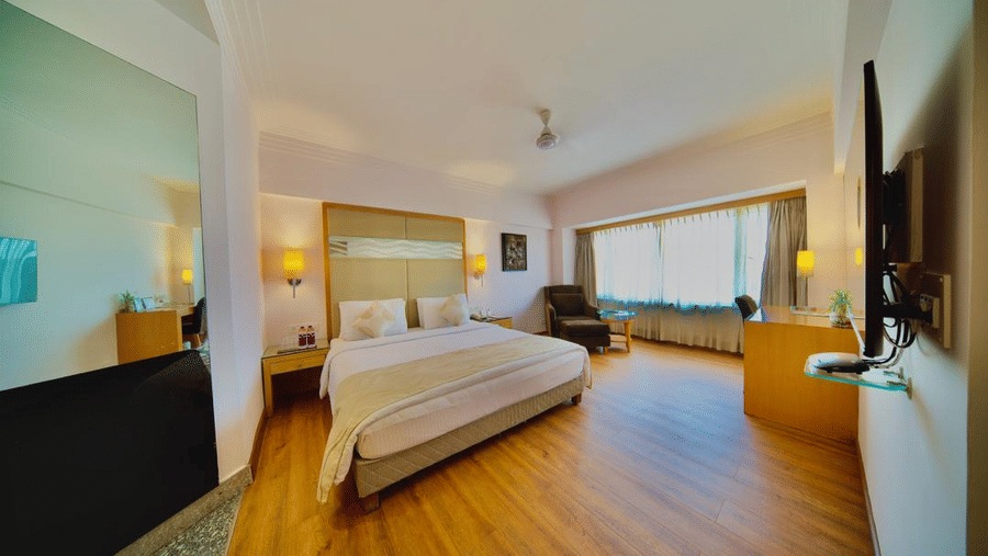 An elegant bedroom in Lords Plaza Ankleshwar with a double bed, wooden panelled floor, large mirror, TV, dim light lamps, and ample natural light from a large window.