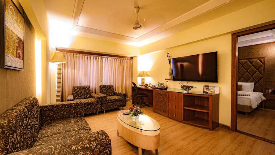 A modern sitting room of a suite in Lords Plaza Ankleshwar with a TV, work desk, large window, cushioned seating, and a flower centrepiece on a table, offering a glimpse of the bedroom
