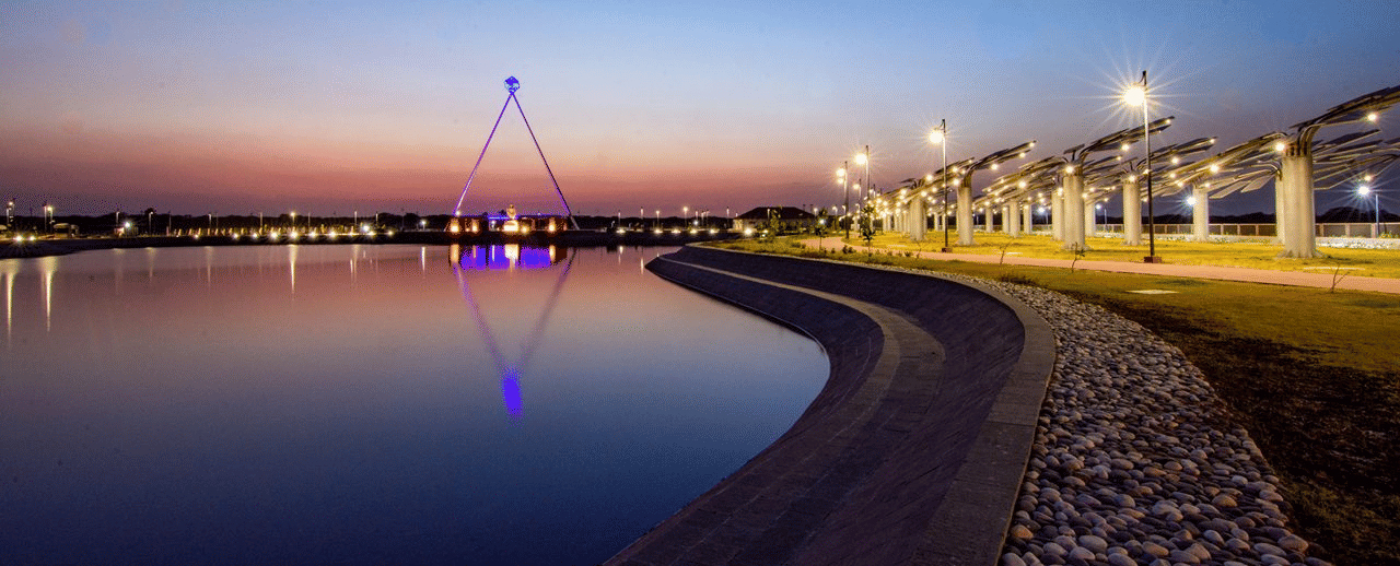 A lake and paved walkway illuminated by streetlights, with a distant cable-stayed tower reflected in the water at twilight