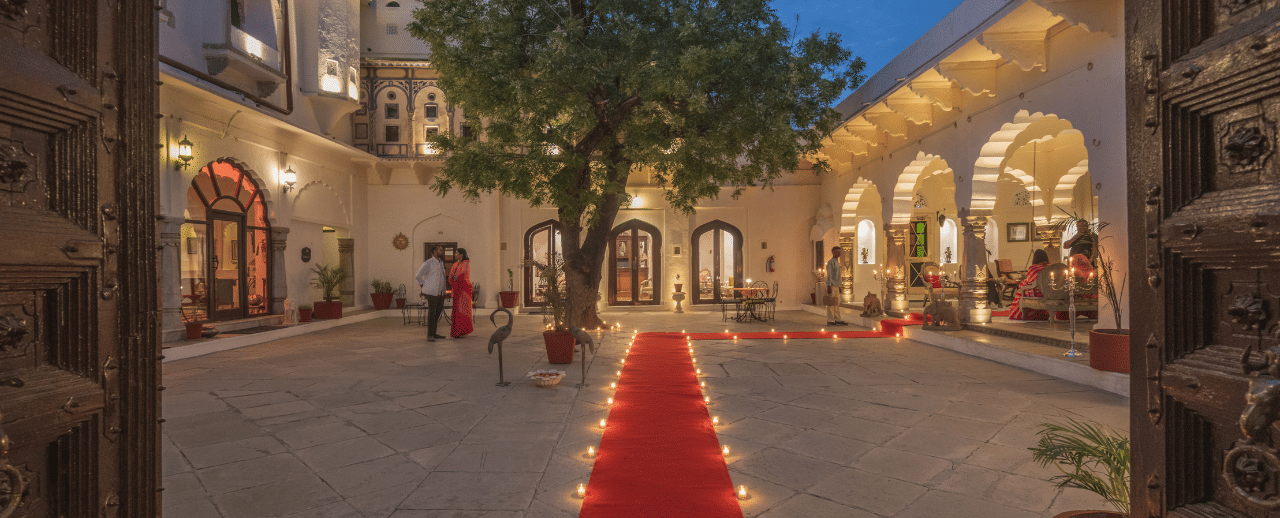 Evening view of a heritage palace courtyard with a red carpet lined with oil lamps leading towards a large tree, surrounded by warmly lit arched facades and guests mingling under the twilight sky.