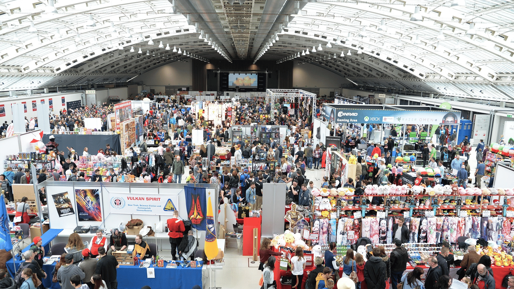 A bustling exhibition hall with a high vaulted ceiling, filled with many stalls and crowds of people at an event.