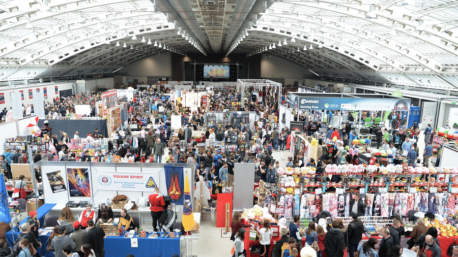 A bustling exhibition hall with a high vaulted ceiling, filled with many stalls and crowds of people at an event.