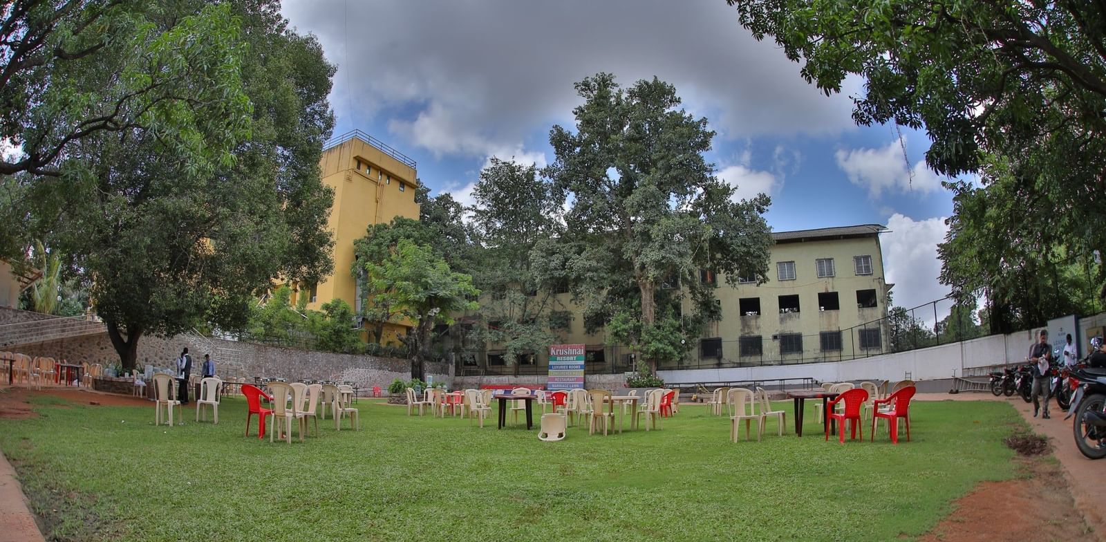 Green lawn at Krushnai Resort with scattered chairs and tables under a cloudy sky, surrounded by tall trees and distant buildings.