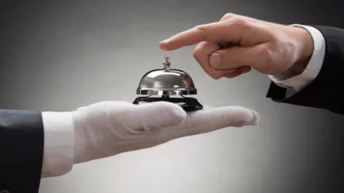 A person in a suite jacket pressing a service bell held on the gloved palm of another person against a grey background.