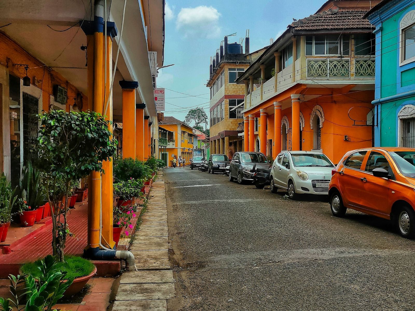 A vibrant street in a heritage town, lined with colourful buildings, potted plants, and parked cars under a bright sky.