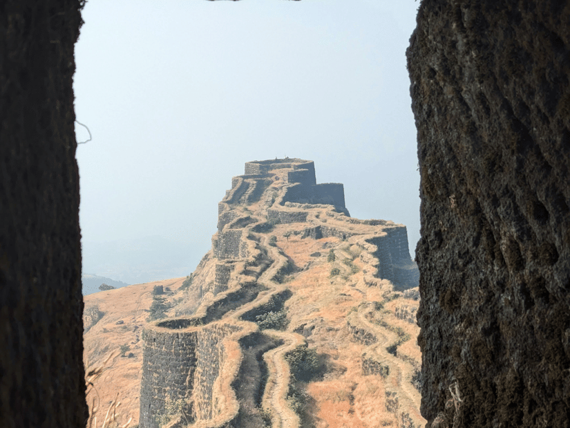 A view framed through a dark stone window or opening, looking out at a stone wall path winding up a rocky hill.