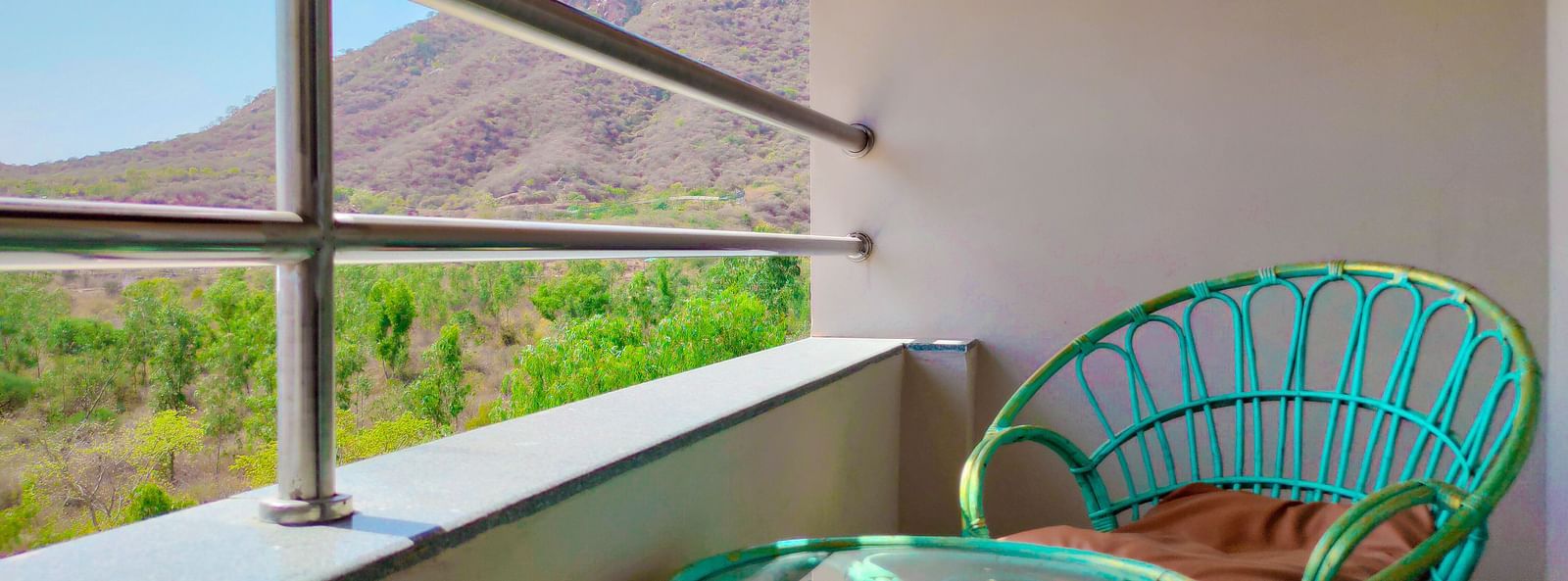 A balcony with a wicker chair and table overlooks a scenic mountain view. - Bamboo Saa Resort & Spa, Udaipur