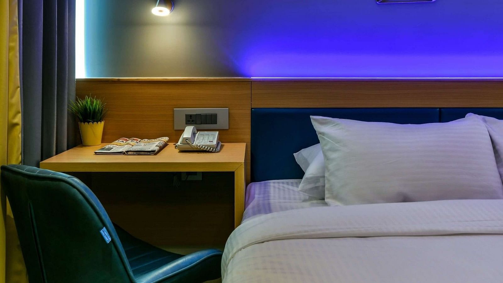 A spacious bed with a wooden work desk and chair placed beside it, accompanied by a night light above, at Indie Stays, BKC Kalina.