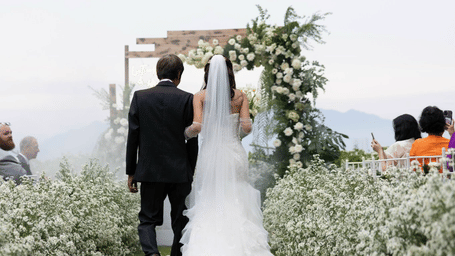 A groom and bride walk down an outdoor aisle towards a floral archway - Alibu