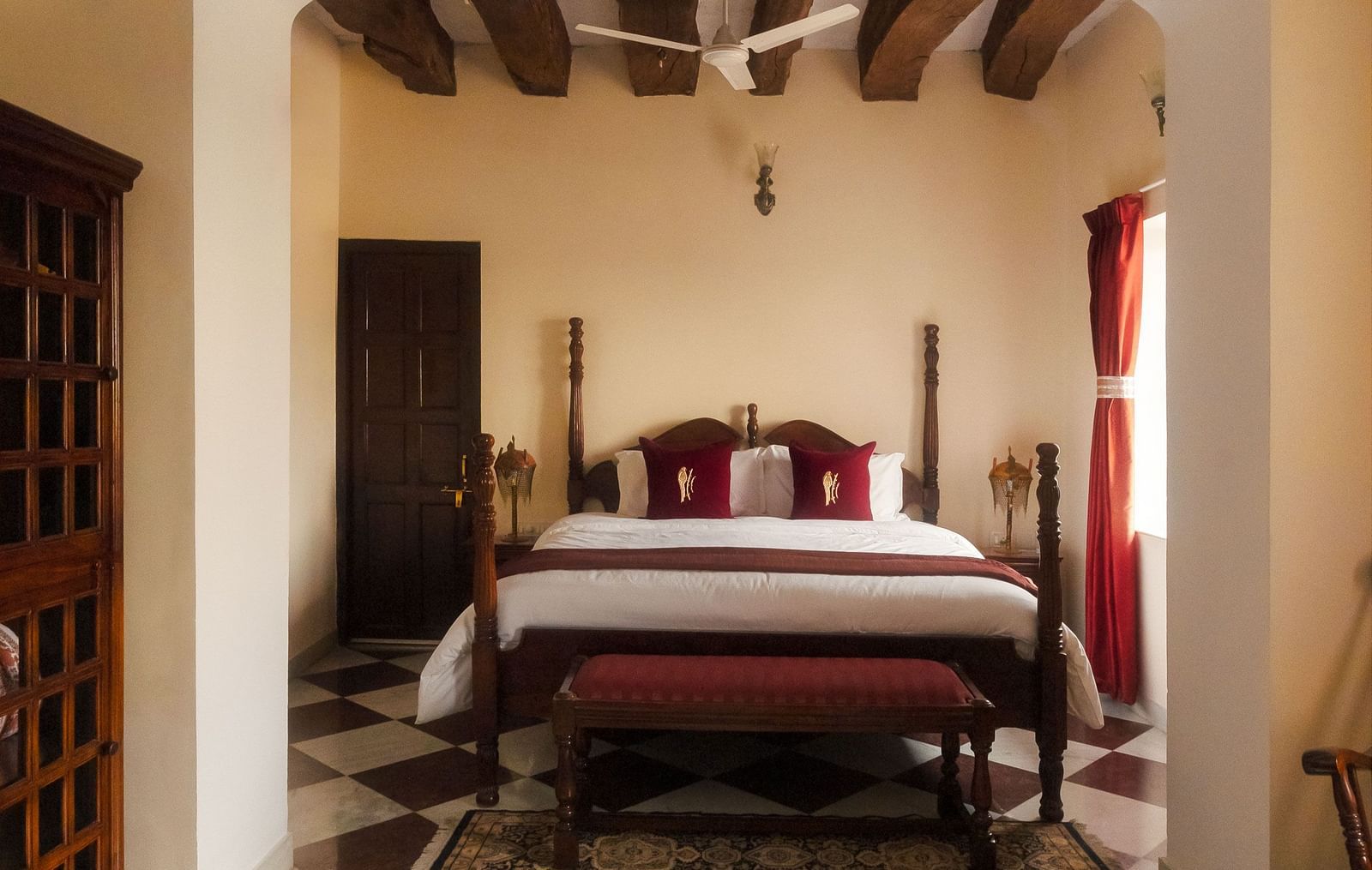 A bedroom at Fort Barli, viewed through an arched doorway, features a four-poster bed, checkered floors, and exposed wooden beams.