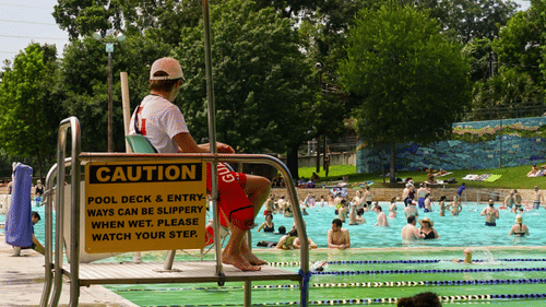 A lifeguard sits on a high stool overlooking the pool, while a woman relaxes at the edge of the swimming pool.