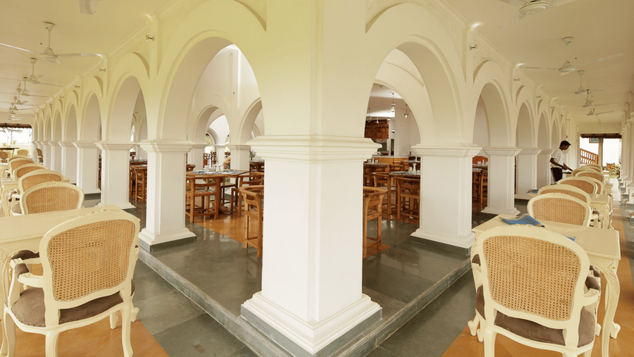 An indoor restaurant dining space featuring white arched pillars and rows of wooden dining tables and chairs | Le Pondy