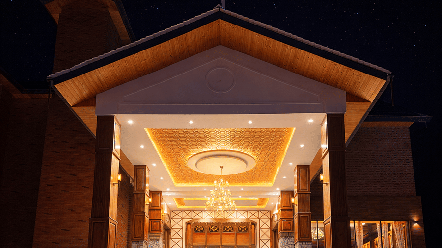 Facade of Mountnoor Sarovar Premiere resort in Pahalgam, with dramatic night view of the main entrance portico. The brightly lit, high-ceilinged entryway features a large chandelier and a central staircase, exuding a warm welcome.