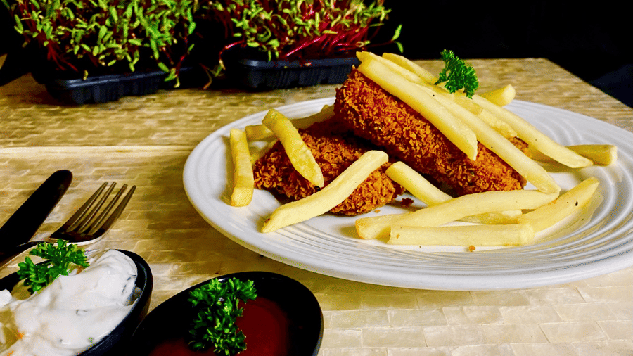 White plate of crumb-fried fish fillets and golden french fries with parsley, served with bowls of tartare sauce and ketchup.