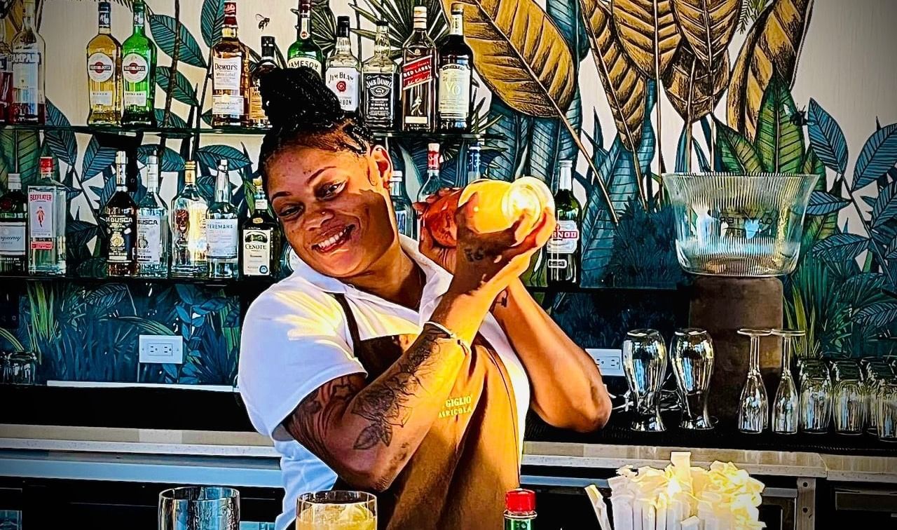 A bartender is shaking a cocktail shaker behind a bar with bottles and tropical-themed decor in the background.