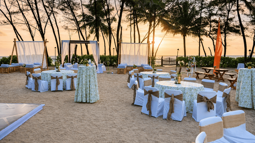 Outdoor beachfront event setup with decorated dining tables under palm trees at sunset at Kenilworth Resort & Spa, Goa.