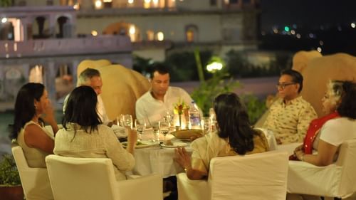 Four people are enjoying an evening meal at a candlelit table outdoors at Tijara Fort-Palace, a Neemrana property in Alwar, with the illuminated fort in the background.