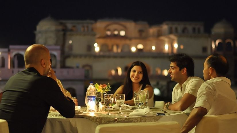 Four people are enjoying an evening meal at a candlelit table outdoors at Tijara Fort-Palace, a Neemrana property in Alwar, with the illuminated fort in the background.