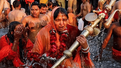 Devotees performing a sacred ritual bath in a river, holding religious symbols amid a dense crowd.