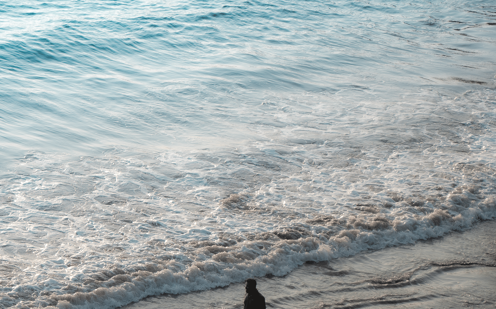 An aerial or high-angle shot of a person walking on a sandy beach next to the foamy edge of light blue ocean waves.