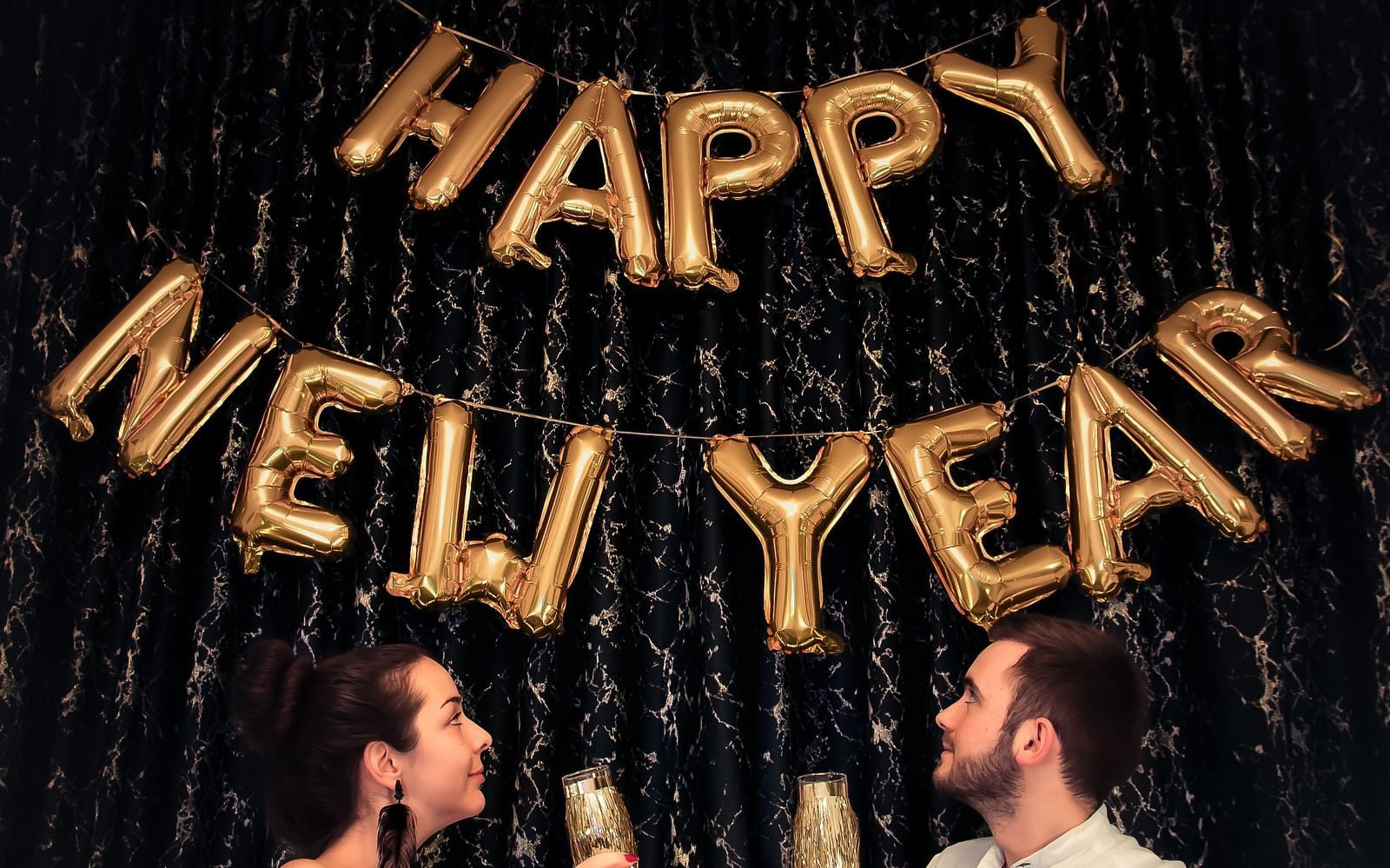 A couple holding drinks with happy new year made out of ballons on the wall