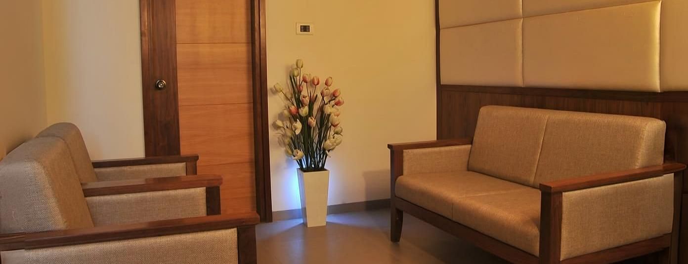 A lounge area with a wooden ceiling, two single-seater chairs, one two-seater sofa, a flower vase placed near a wooden door, and softly lit recessed lights on the ceiling.