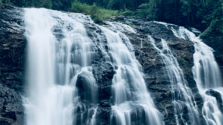 A wide Abbi Gundi Falls cascades over dark rock into a green pool, surrounded by dense forest and towering trees beneath an overcast sky.