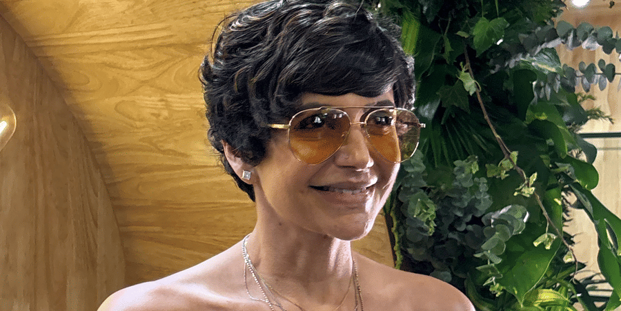 Woman with a short pixie haircut and tinted sunglasses wearing a white strapless top with black polka dots in front of a wooden wall with neon signage.