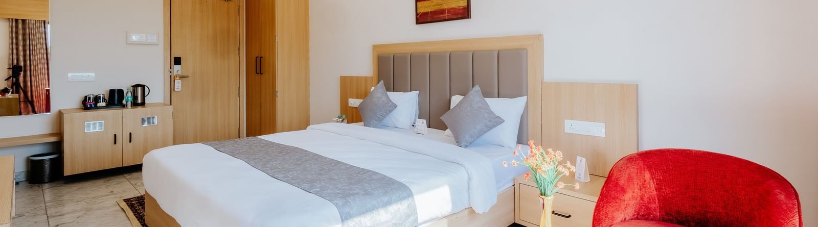 Corner view of a brightly lit bedroom at Daksh The Grand Gowardhan, Dwarka, showcasing a plush double bed, a vibrant red armchair along with a coffee table in front of it.