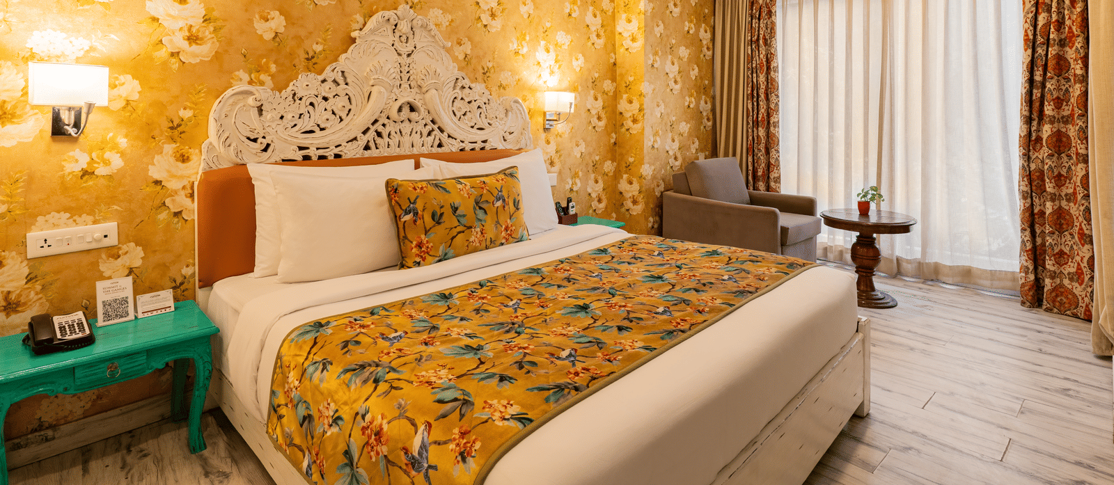 A spacious bedroom featuring a bed with a decorated headboard, an accent wall with floral wallpaper, a bedside table, and a single-seater sofa facing a small round table | Summit by the Ganges Beach Resort & Spa, Rishikesh