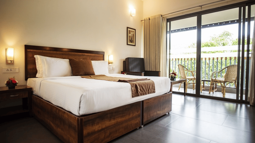 Bright bedroom at Abad Green Forest, Thekkady with wooden furnishings, crisp white bedding, and sliding doors opening to a balcony overlooking greenery.