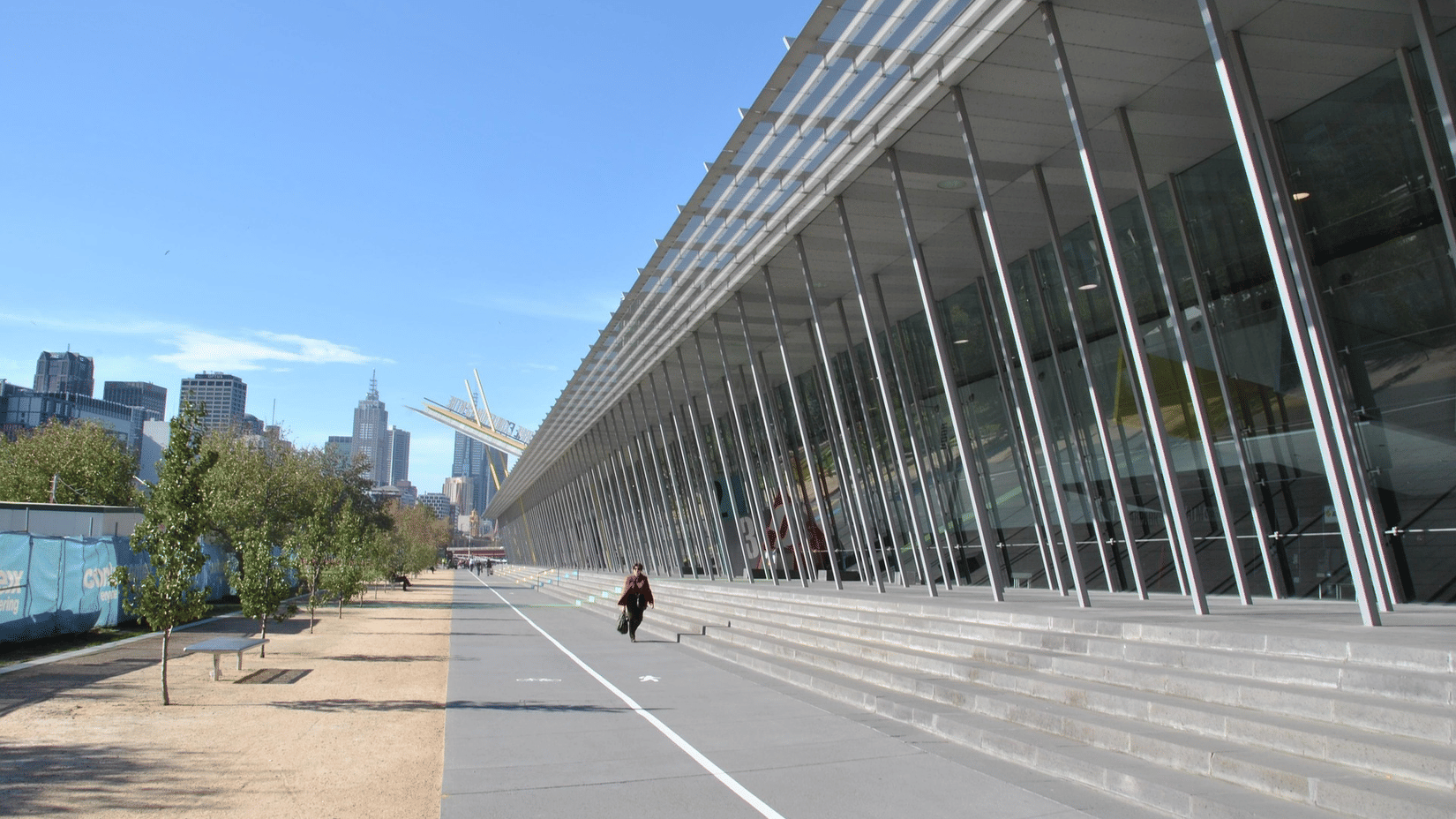 Exterior of a long, modern glass building with a concrete pavement and city skyline under a clear blue sky.