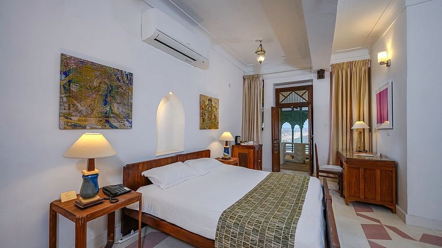 Room with bed, wooden frame, patterned runner, furniture including tables and cabinets, lamps, artwork, curtains, and doorway leading to another room - Tijara Fort-Palace - 19th Century, Alwar.