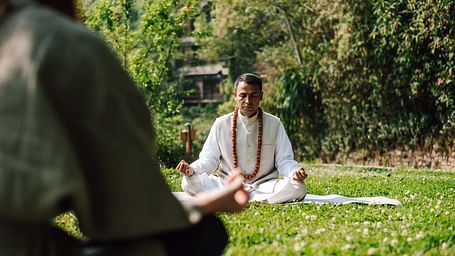 Person meditating outdoors in a peaceful garden setting, surrounded by greenery.