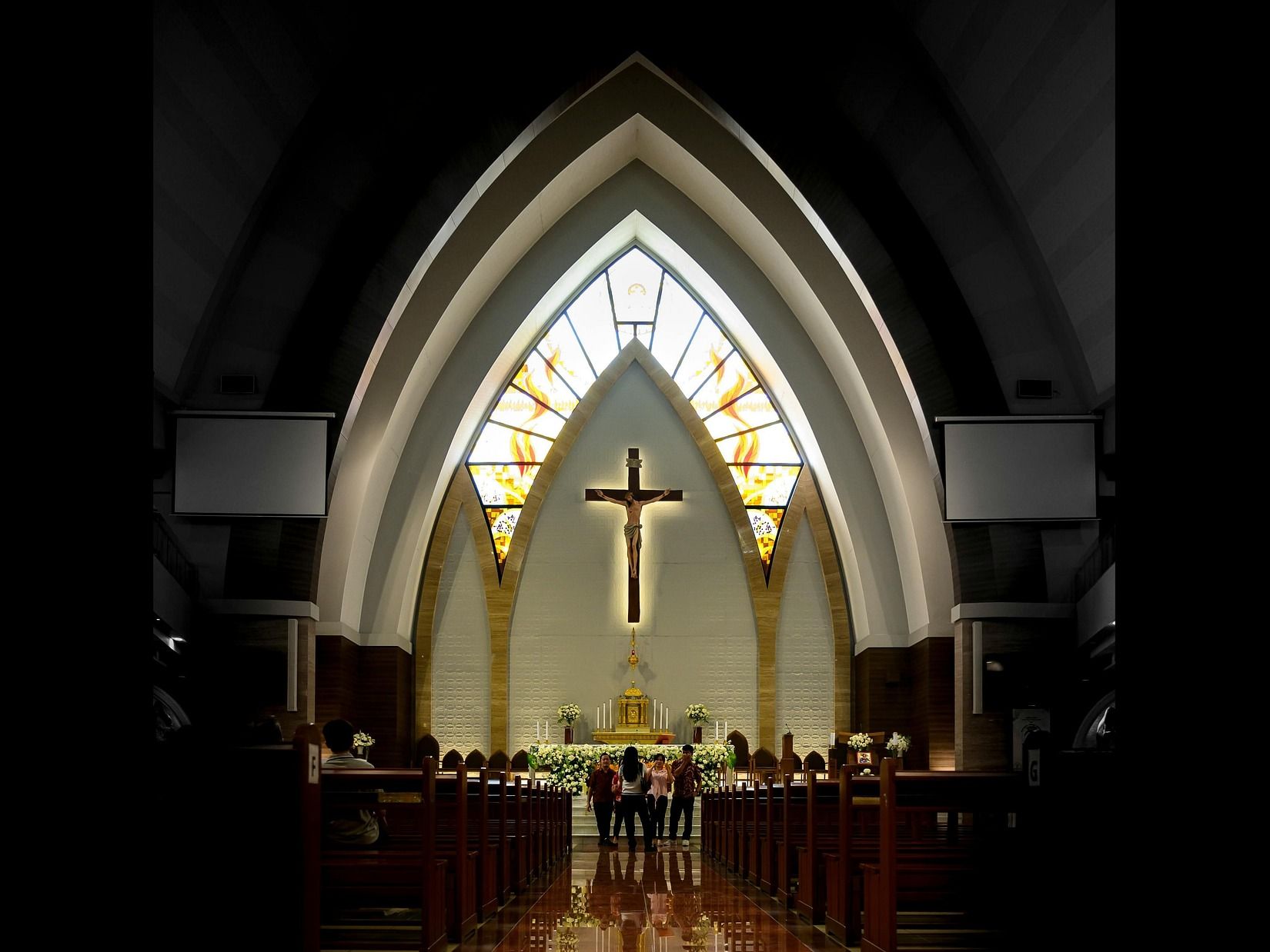 The interior of a church features a central aisle leading to an altar with a cross, stained glass windows, and rows of pews with people.