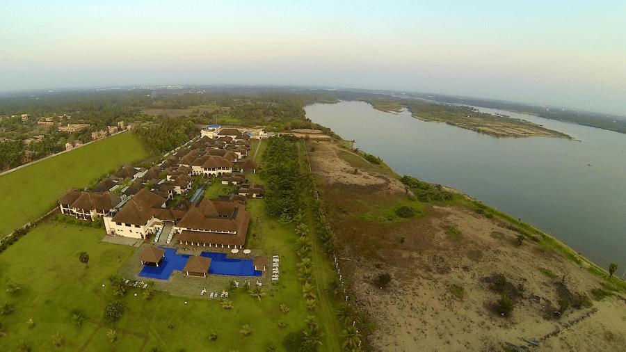 A wide aerial view of the resort property showing the buildings, swimming pool area, and the adjacent river under a clear sky | Le Pondy