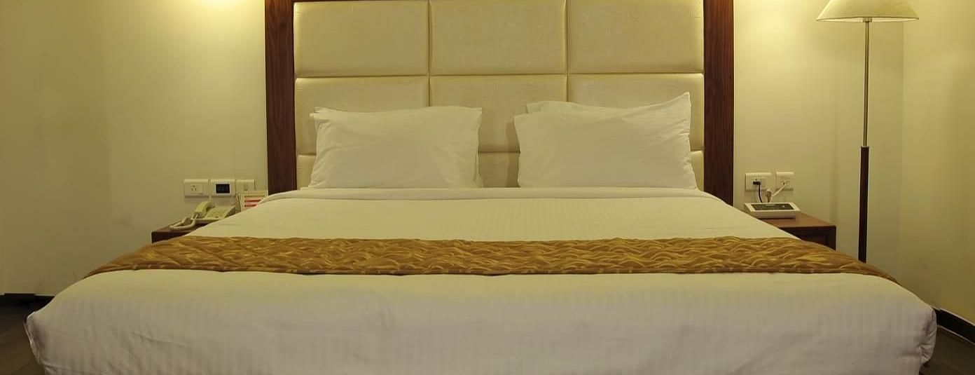 A queen size bed with white linens and a gold runner, two hanging lights, a floor lamp, and a white rug on a wooden floor at Dhanalakshmi Srinivasan Hotels, Perambalur.