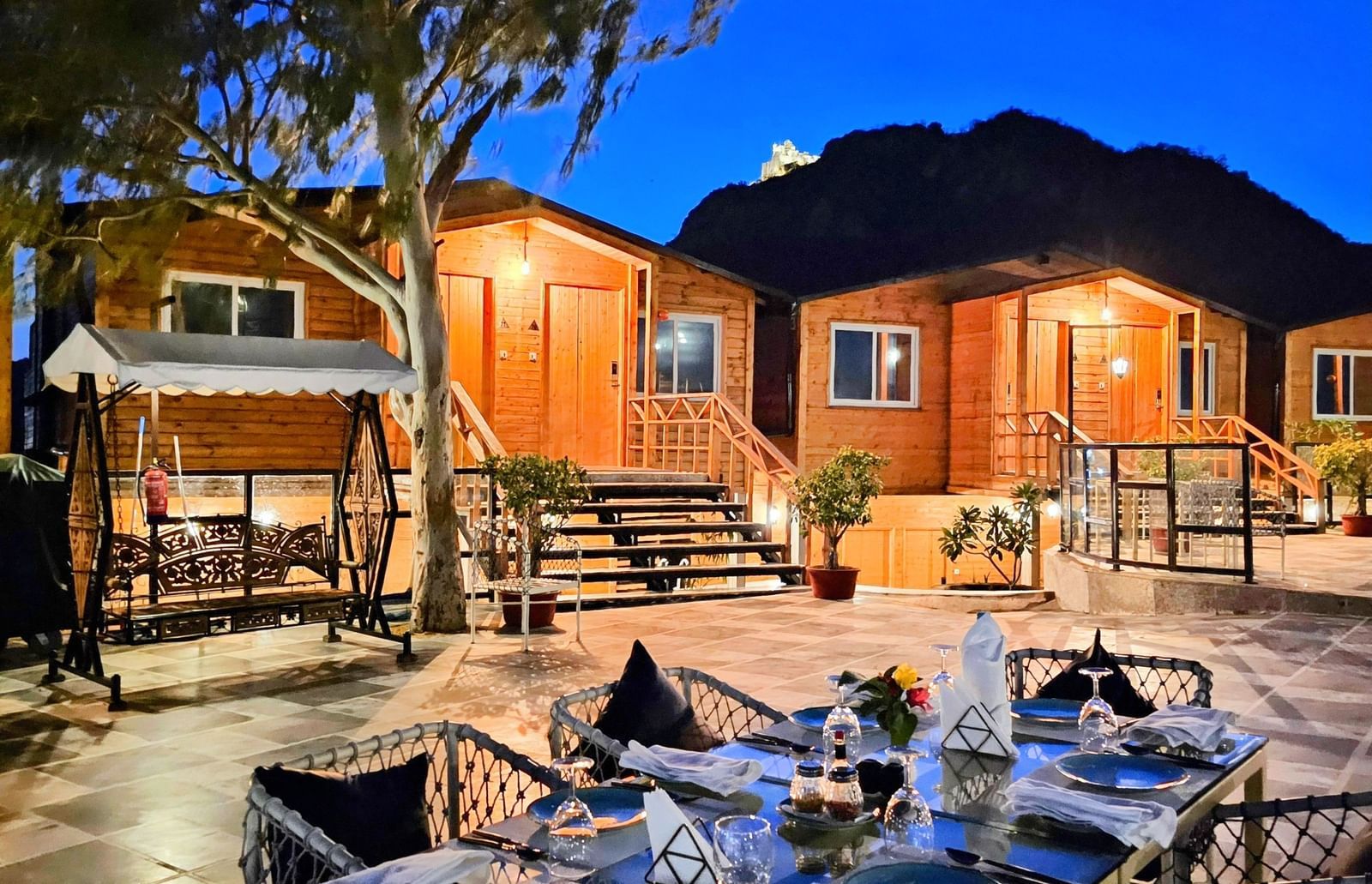 An outdoor dining table with wooden cabins in the background. - Bamboo Saa Resort & Spa, Udaipur.